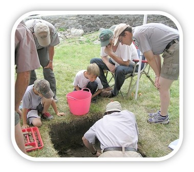 Archaeologist at work on dig at Colonial Pemaquid State Historic Site in Maine, with onlookers standing over dig hole.