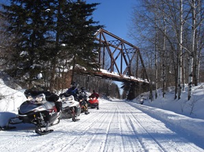 Three snowmobiles sit on the side of a snow-covered trail under a bridge.