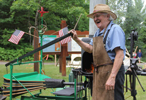 A blacksmith stands in front of his furnace, arm resting on the handle to his bellows, and laughs