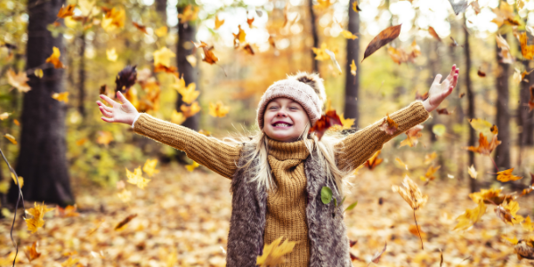 Young girl with warm clothes and wooly hat. She is standing in an autumn forest with leaves falling. She is smiling.