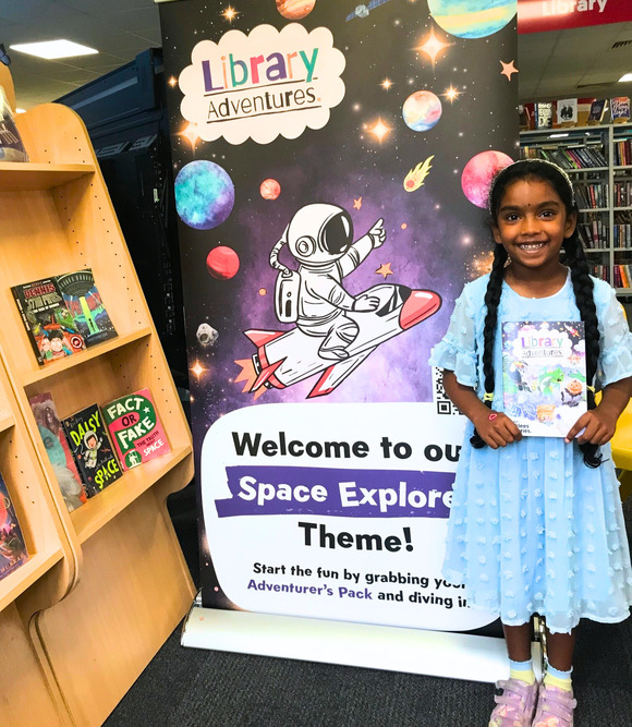 Girl in blue dress with beautiful smile holding Library Adventures booklet in Huddersfield Library