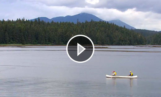 Video still of two people rowing a boat on a lake with mountains in the background.