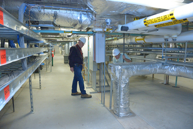 Two men examine exhaust pipe in floor of building filled with mechanical tracking, pipes and control boxes.