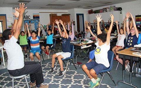 Students in classroom sitting at their desks with their arms up