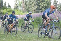 Cyclists race through a flat grassy spot next to a field of fireweed