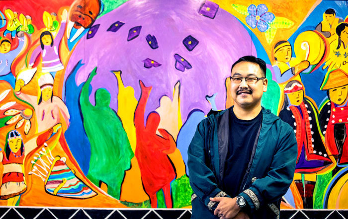 Percy Avugiak stands in front of a mural he painted at UAF’s Rasmuson Library entitled Invitation Dance and Celebrations.