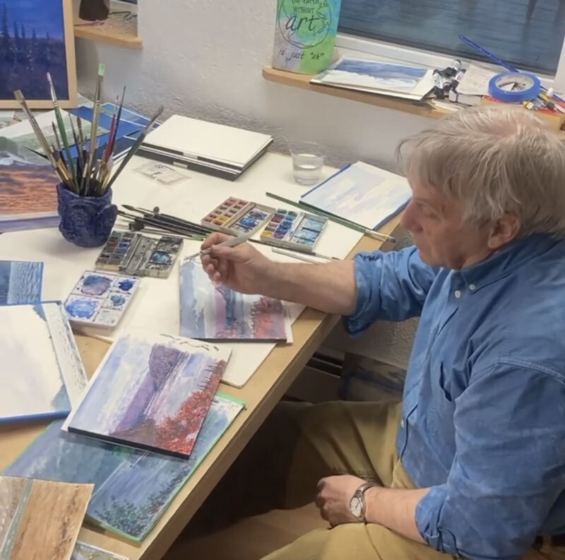 David Rosenthal sits at a table full of watercolor palettes and brushes