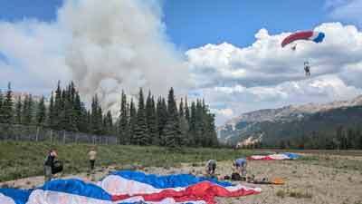 A smokejumper descends to the ground via parachute, while four others prepare their gear on the ground. 