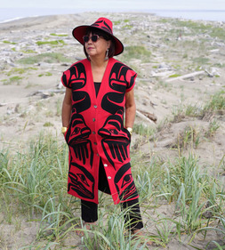 Dorothy Grant in red and black formline sleeveless tunic and hat with grasses, sand, water in background