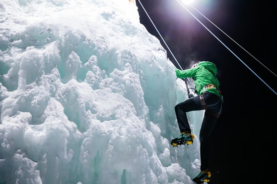 An climber scales a wall of ice in the moonlight wearing a harness and climbing gear.