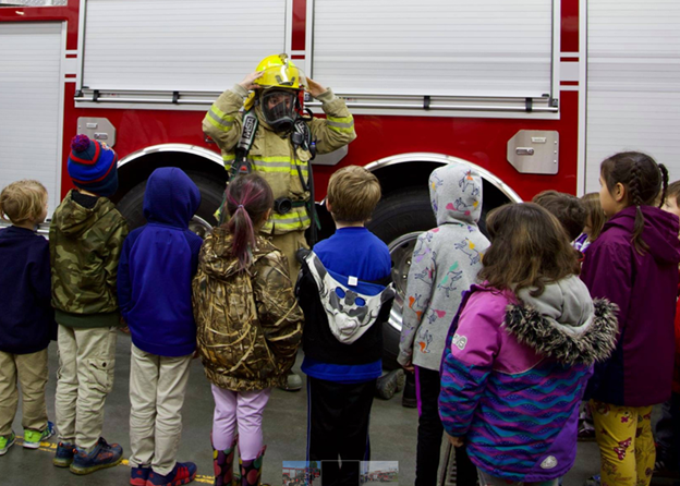 A firefighter points to their helmet and explains to a group of children how this piece of equiptment protects them in a fire.