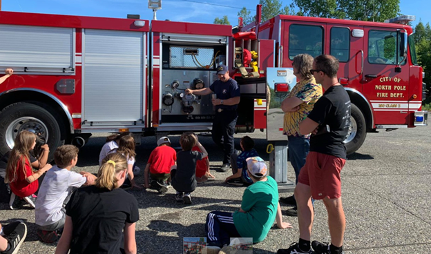 A group of children sit near a fire engine while a firefighter shows them the equipment and talks about fighting fires.