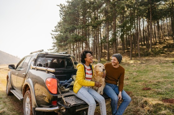 Two people sit on the tailgate of a truck enjoying the outdoors with their dog.