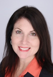 Professional headshot of Rebecca Cowan. She is wearing a dark blazer over an orange collared shirt, with a light gray background.