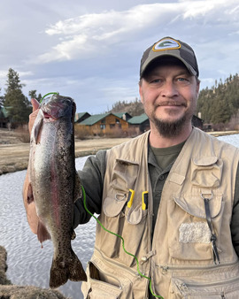 Brian N man holding large rainbow trout standing in front of a pond with some winter trees in the back