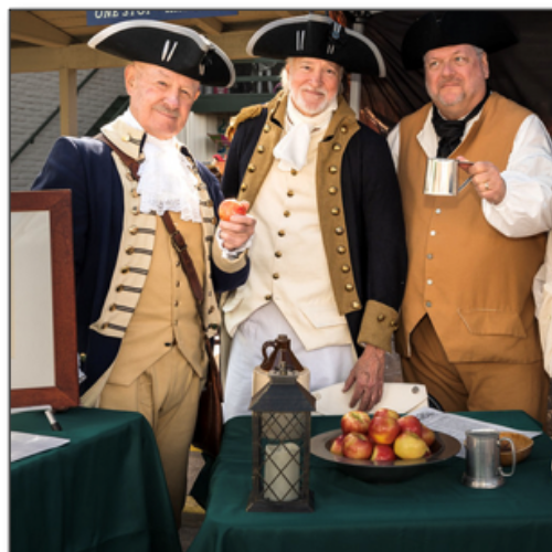 Four people in colonial attire stand behind tables displaying apples, a lantern, and pewter mugs.