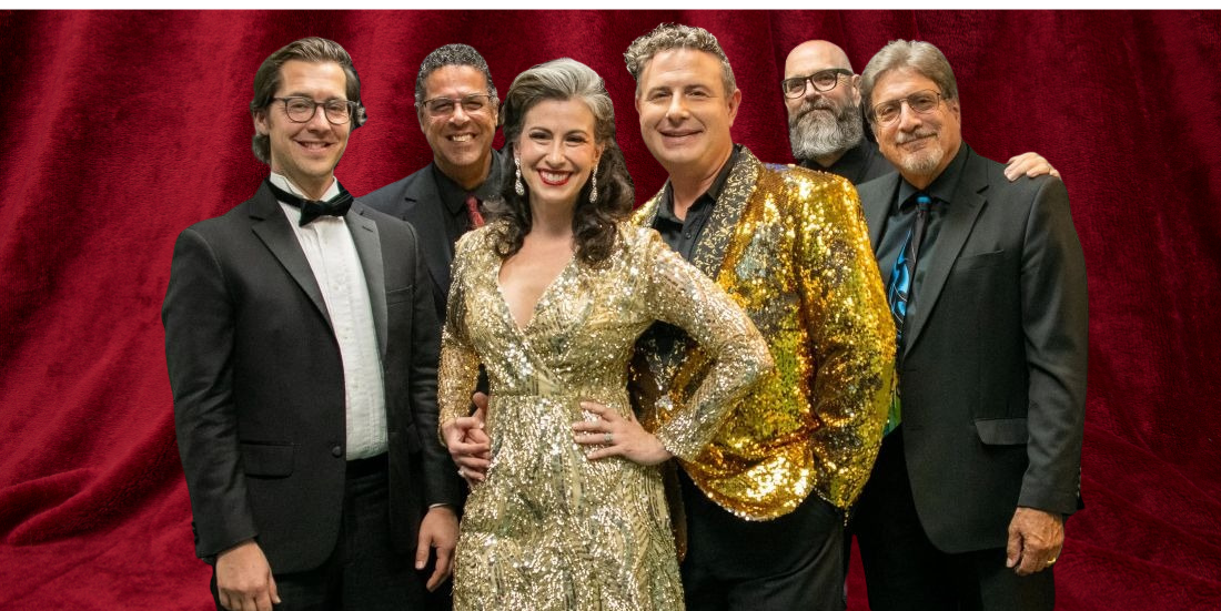 A group of six people, five men and one woman, pose for a photo. The woman in the center wears a sparkling gold dress.