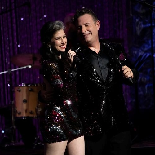 A woman in a sequined dress embraces a man in a matching jacket.  Both hold microphones and are facing an audience. 