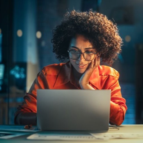 A black woman works at a laptop with an awed expression against a nighttime backdrop. 