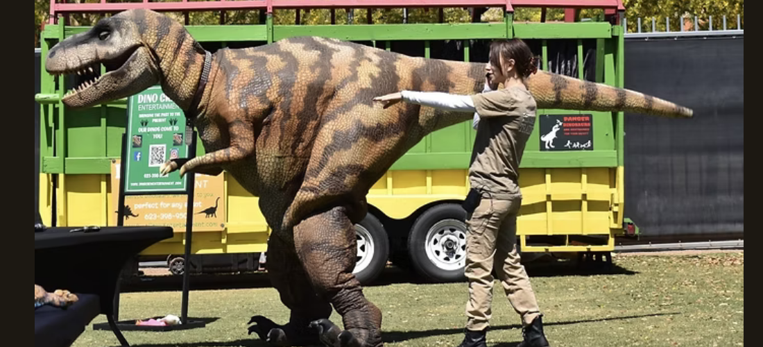 A person in khaki clothing operates a dinosaur costume in front of a trailer with a dinosaur graphic.