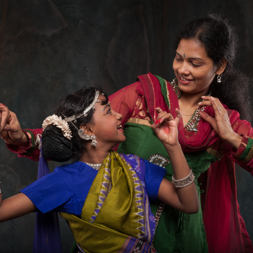 Two dancers in traditional Indian attire, one younger and one older, look at each other and smile, adorned with jewelry and colorful fabrics.