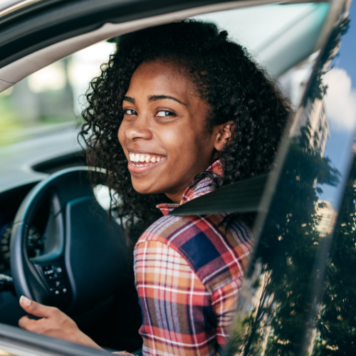 A woman with curly hair wearing a plaid shirt smiles as she sits in the driver's seat of a car.