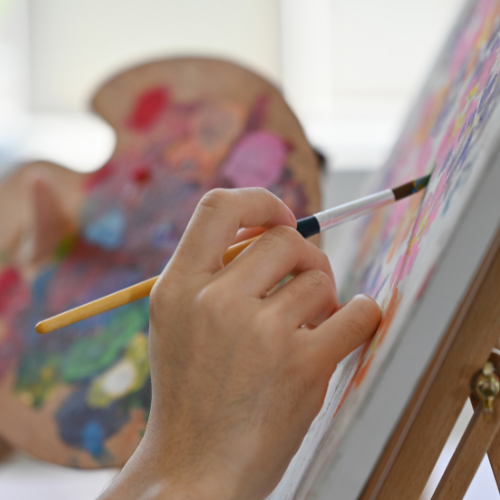 A hand holding a paintbrush and applying paint to a canvas on an easel, with a colorful palette in the background.