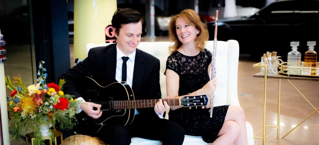 A man in a suit plays a guitar while a woman in a black dress holds a flute, seated next to a floral arrangement and a bar cart with decanters.