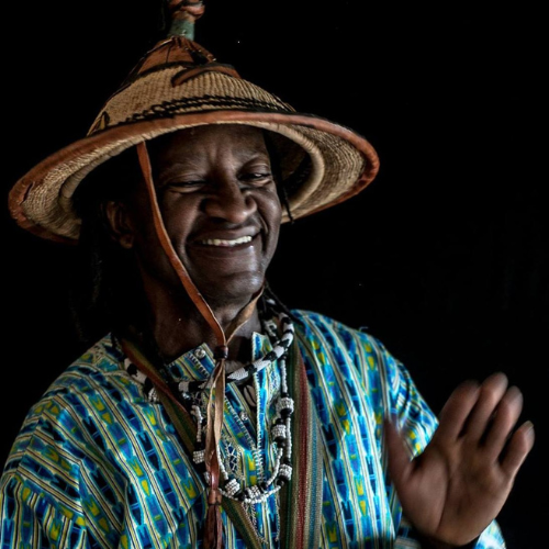 A person wearing a woven conical hat and a blue and green patterned shirt smiles warmly, with beads around their neck.