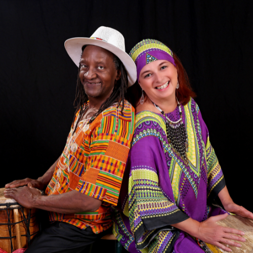 A man in a kente cloth shirt and white hat and a woman in a purple dashiki and headscarf sit back-to-back with drums in front of them.
