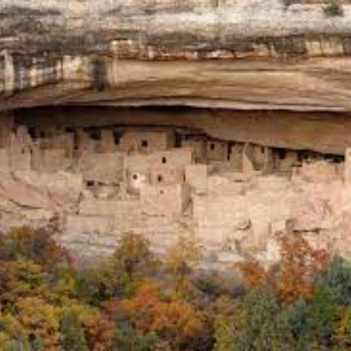 Ancient cliff dwellings built into a rock overhang, with trees displaying autumn colors in the foreground.