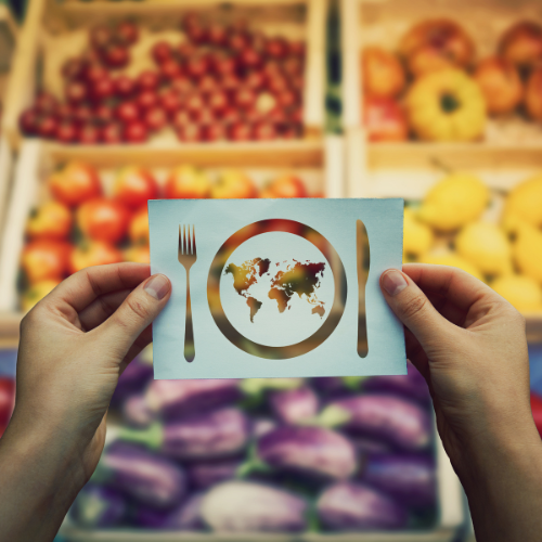 A person holds a card with a world map on a plate, with a fork and knife, in front of a blurred background of fresh produce.