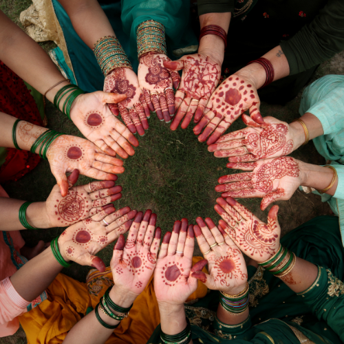 A circle of hands adorned with intricate henna designs and green bangles, viewed from above.