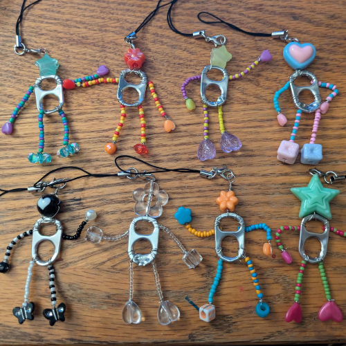 A collection of seven handmade charms resembling people, crafted from soda can tabs and colorful beads, arranged on a wooden surface.