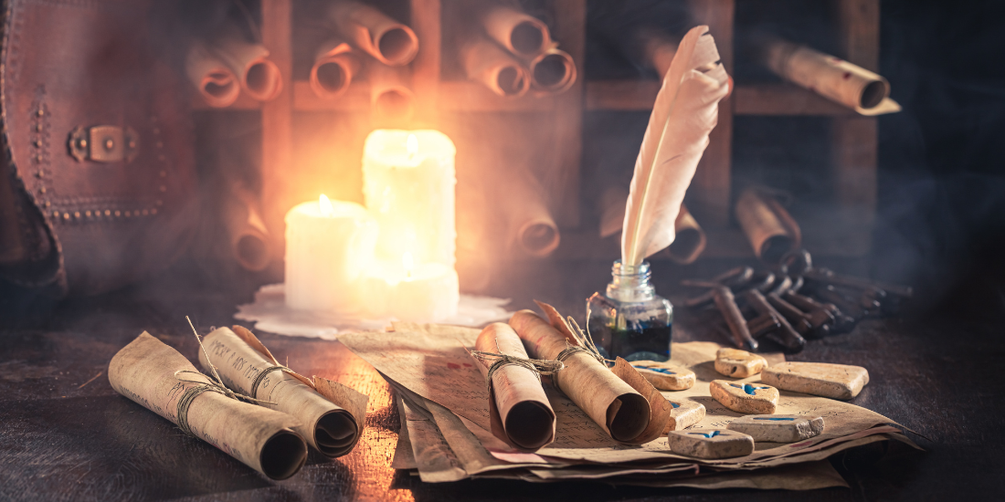 A quill pen rests in an inkwell next to rolled scrolls and lit candles on a wooden desk.