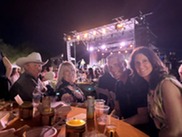 Group of four people sitting at table with stage and lights in background