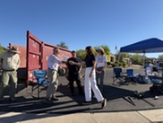 Councilmember shaking resident's hand in front of large red dumpster and blue tent (outside)