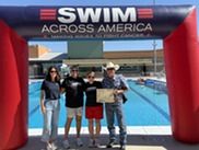 People standing in front of pool and Swim Across America Sign