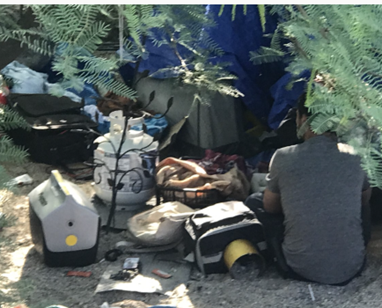 Homeless man sitting under brush next to his belongings and garbage