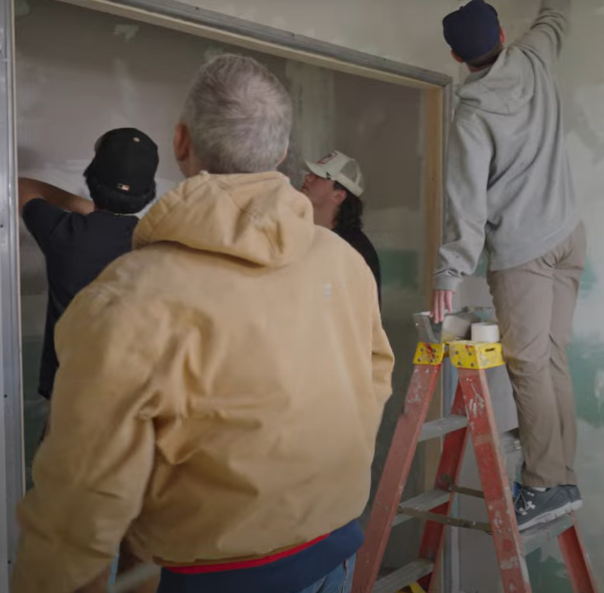 Picture shows 3 students paint the tiny home wall supervised by the contractor
