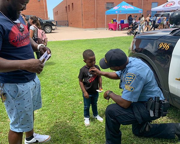 A male TPD CSO kneels down to put a badge sticker on a little boy while an adult man looks on, smiling.