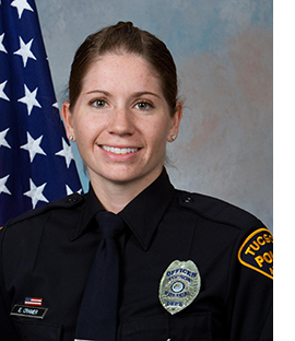 Department photo of smiling female officer in uniform in front of an American flag.