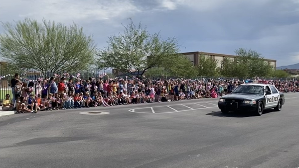 art of a parade, a TPD patrol vehicle drives slowly past assembled students outside a school.