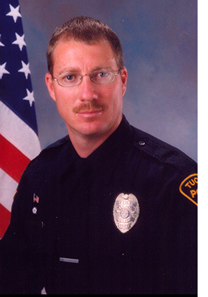 Department photo of a TPD male officer in uniform in front of an American flag.