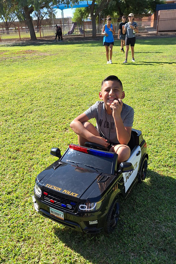 Smiling boy sits in a mini toy TPD patrol vehicle on grass, with people walking in the background.