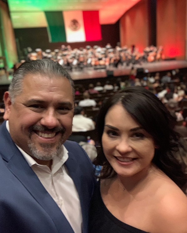 Hispanic TPD Lt and his wife indoors at a festive event with a large Mexican flag in the background.