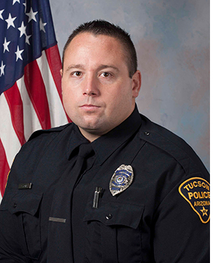 Department photo of male officer in uniform in front of an American flag.