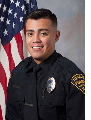 Department photo of a smiling male officer in uniform in front of an American flag.