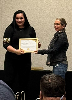 Two women are standing. One hands the other a framed certificate.