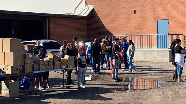 Cardboard boxes containing food are spread out on a table outside, with volunteers behind.